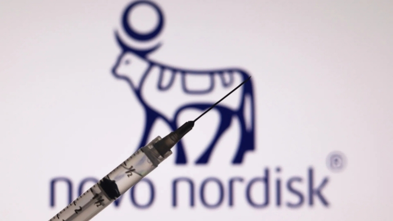 Syringe in foreground with blurred Novo Nordisk logo behind