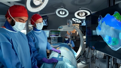 Two surgeons in red caps under operating theatre lights with a digital anatomical model displayed on a monitor